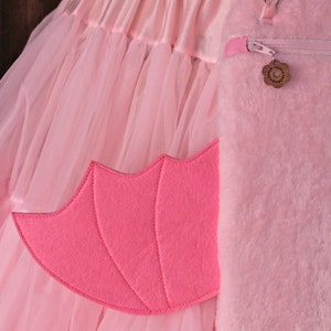 May include: A pink tulle skirt with a pink furry bag attached. The bag has a zipper and a small pink flower charm. The skirt has a pink felt wing attached to the side.