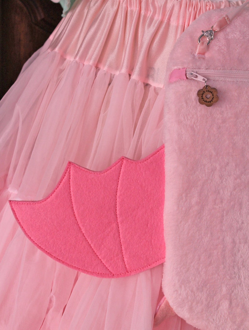 May include: A pink tulle skirt with a pink furry bag attached. The bag has a zipper and a small pink flower charm. The skirt has a pink felt wing attached to the side.