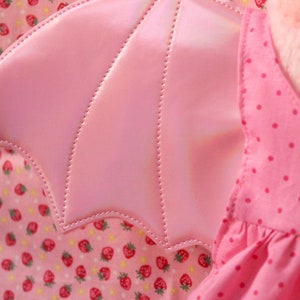 May include: Pink fabric wings with a scalloped edge and a stitched design. The wings are attached to a pink dress with white polka dots.