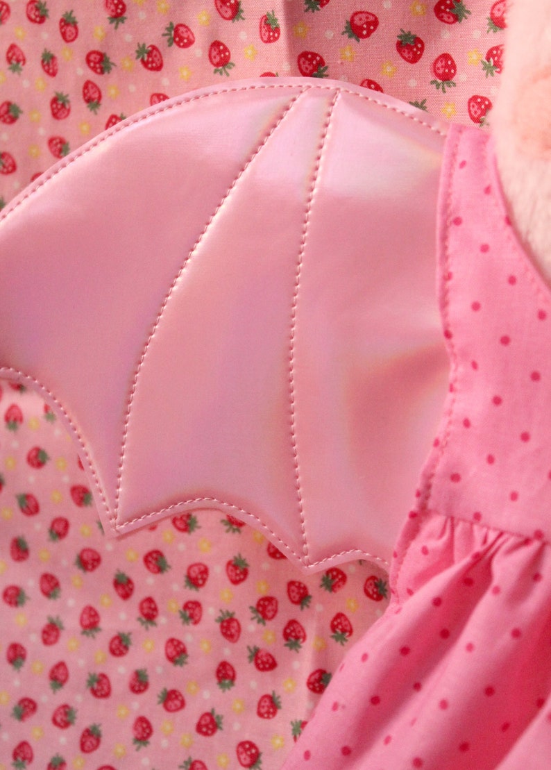 May include: Pink fabric wings with a scalloped edge and a stitched design. The wings are attached to a pink dress with white polka dots.