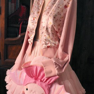 May include: A pink satin bomber jacket with floral print, worn over a pink tulle skirt and a pink axolotl plush bag with iridescent wings.