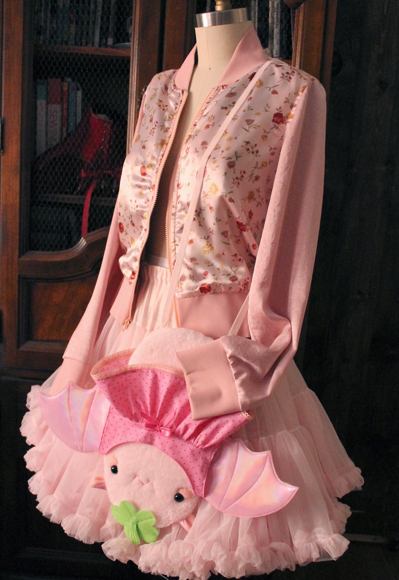 May include: A pink satin bomber jacket with floral print, worn over a pink tulle skirt and a pink axolotl plush bag with iridescent wings.