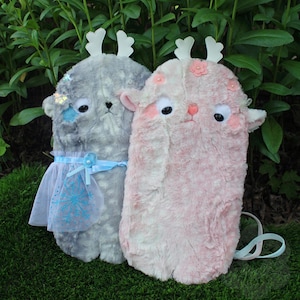 May include: Two plush, furry creature-shaped backpacks. One is gray with a blue skirt and snowflake detail, the other is pink with flower accents. Both have white antler-like details and large, black eyes.