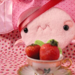 May include: A pink plush toy wearing a pink and white polka dot dress with a red ribbon. The toy is holding a teacup with two red strawberries inside.