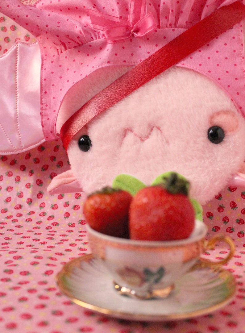 May include: A pink plush toy wearing a pink and white polka dot dress with a red ribbon. The toy is holding a teacup with two red strawberries inside.