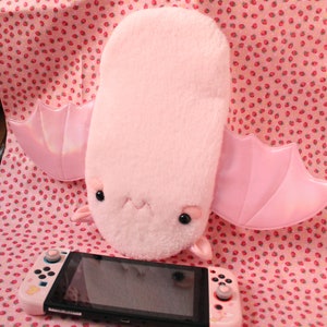 May include: A pink plush bat with large, iridescent wings. The bat is sitting on a pink and white patterned fabric with a Nintendo Switch console in front of it.