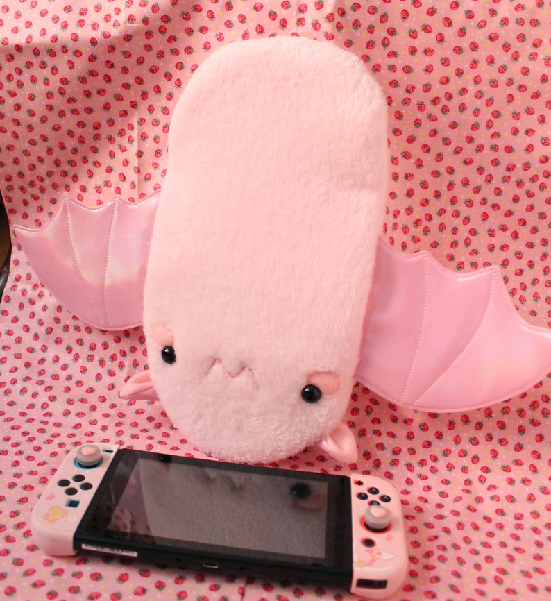 May include: A pink plush bat with large, iridescent wings. The bat is sitting on a pink and white patterned fabric with a Nintendo Switch console in front of it.