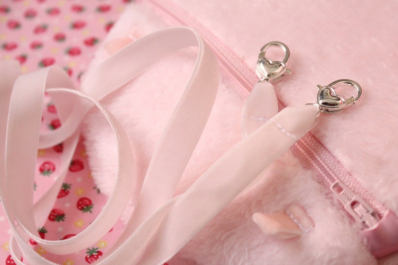 May include: A pink plush bag with a zipper closure and a pink satin strap with silver heart-shaped clasps.