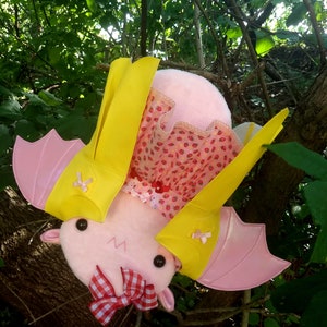 May include: A pink plush bat with yellow wings and a red and white gingham bow. The bat is wearing a pink and white dress with a strawberry pattern.