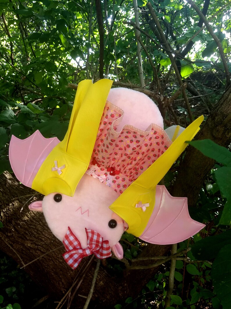 May include: A pink plush bat with yellow wings and a red and white gingham bow. The bat is wearing a pink and white dress with a strawberry pattern.