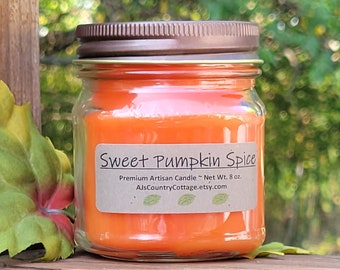 May include: A glass jar candle with an orange wax fill and a brown metal lid. The label reads "Sweet Pumpkin Spice" and "Premium Artisan Candle ~ Net Wt. 8 oz.". The candle sits on a wooden surface with green foliage in the background.