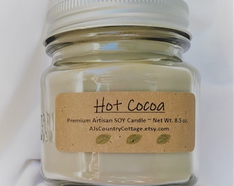 May include: A glass jar candle with a brown label that reads "Hot Cocoa" and "Premium Artisan SOY Candle ~ Net Wt. 8.5 oz.  AJsCountryCottage.etsy.com".