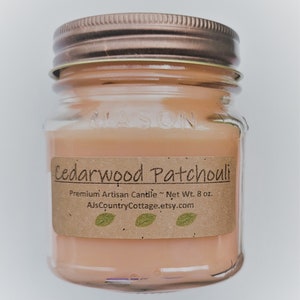 May include: A clear glass jar candle with a brown lid and a brown paper label. The label reads "Cedarwood Patchouli", "Premium Artisan Candle", "Net Wt. 8 oz.", and "AlsCountryCottage.etsy.com".