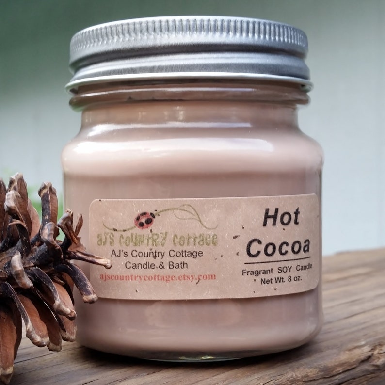 Puede incluir: Una vela de vidrio con una mezcla de cera de soja marr&oacute;n. La etiqueta dice "Hot Cocoa" y "Fragrant SOY Candle, Net Wt. 8 oz."