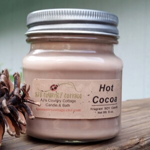 Puede incluir: Una vela de vidrio en un frasco con tapa plateada. La vela es de color marr&oacute;n claro y tiene una etiqueta que dice "Hot Cocoa" y "Fragrant SOY Candle, Net Wt. 8 oz."