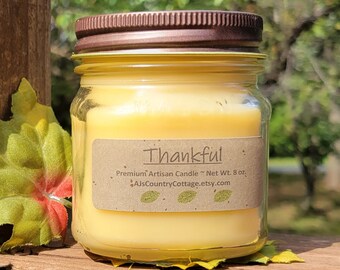 May include: A glass jar candle with a brown lid and a label that reads "Thankful." The candle has a yellow wax and is labeled as a premium artisan candle, net weight 8 oz. The candle sits on a wooden surface with fall foliage.