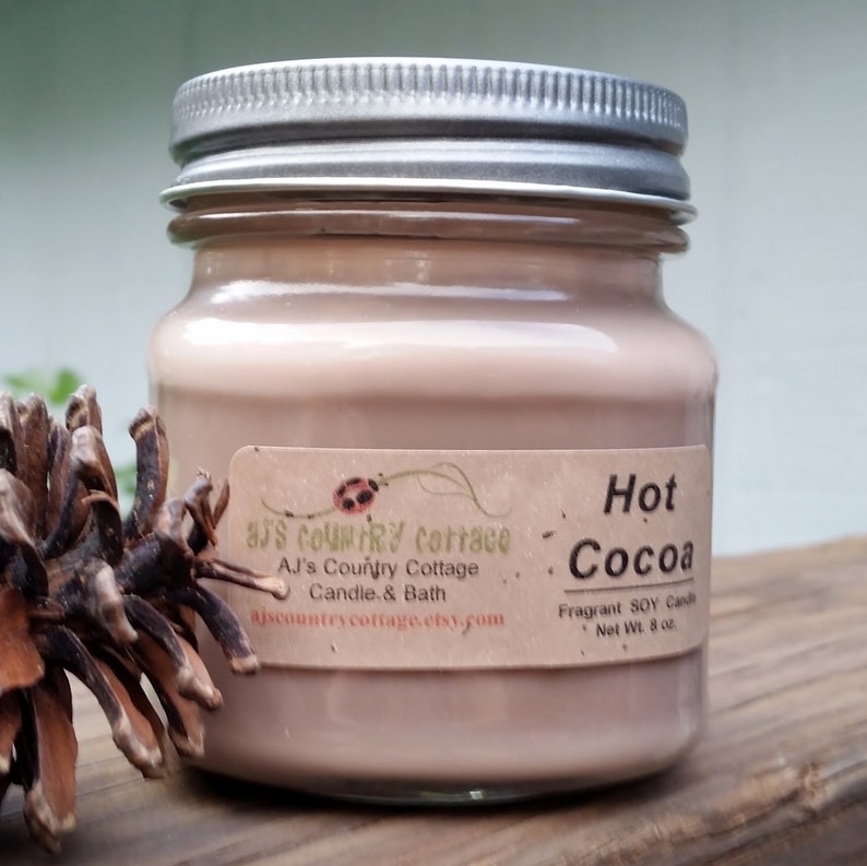 Puede incluir: Una vela de vidrio en un frasco con una tapa plateada. La vela es de color marr&oacute;n claro y tiene una etiqueta que dice "Hot Cocoa" y "AJ's Country Cottage Candle & Bath". La vela pesa 8 onzas.