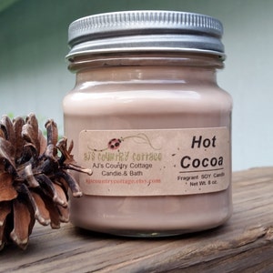 Puede incluir: Una vela de vidrio en un frasco con tapa plateada. La vela es de color marr&oacute;n claro y tiene una etiqueta que dice "Hot Cocoa" y "Fragrant SOY Candle, Net Wt. 8 oz."