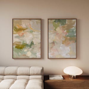 May include: Two framed abstract paintings with gold frames. The artwork features brushstrokes in shades of green, peach, white, and beige. The paintings are displayed on a neutral-coloured wall, creating a modern and artistic aesthetic.