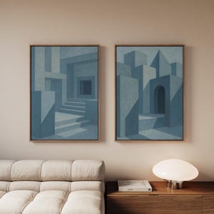 May include: Two framed abstract paintings with a geometric architectural theme. The artwork features blue-grey structures with shadows, set against a light beige background. The frames are a warm, light brown colour.