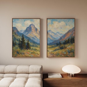 May include: Two framed landscape paintings showing mountain scenes. The artwork uses blues, greens, and browns, focusing on mountains and trees. The paintings are displayed on a neutral wall, with a sofa and lamp in the foreground.