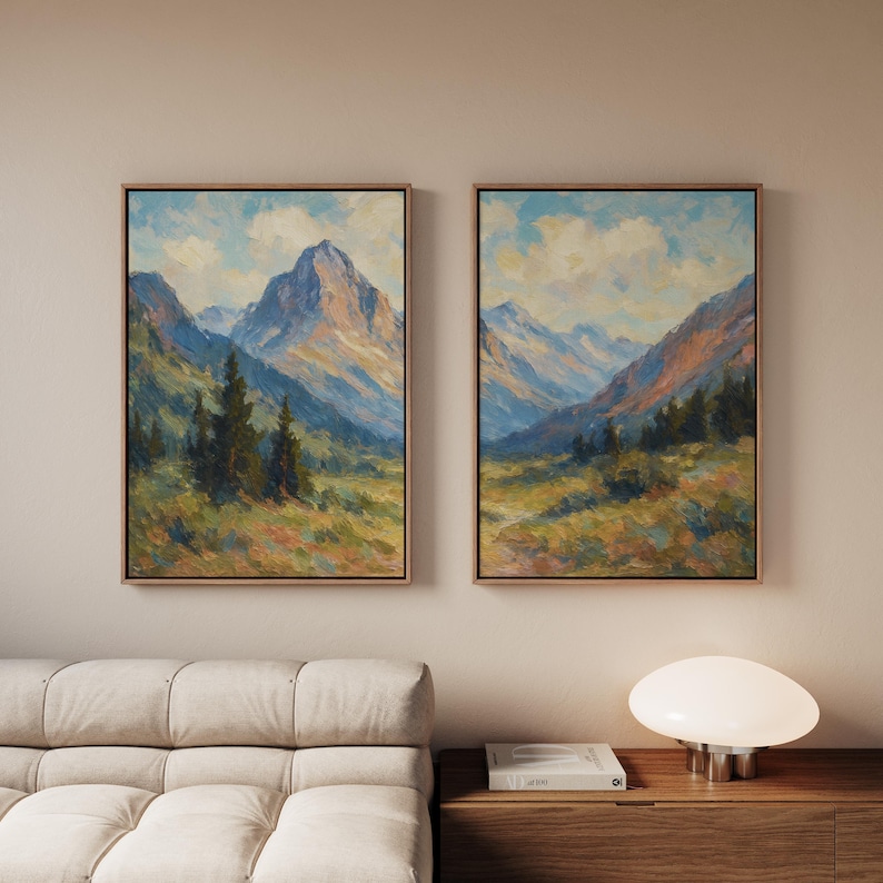 May include: Two framed landscape paintings showing mountain scenes. The artwork uses blues, greens, and browns, focusing on mountains and trees. The paintings are displayed on a neutral wall, with a sofa and lamp in the foreground.