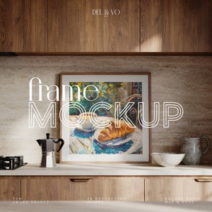 May include: A square frame mockup featuring a painting of a croissant and coffee on a table in a garden setting. The frame is on a countertop in a kitchen with wood cabinets and a light brown backsplash.