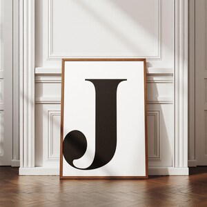 May include: A framed art print featuring a large black letter "J" on a white background. The artwork is displayed in a brown wooden frame and leans against a white wall with decorative molding. The floor is made of dark wood.