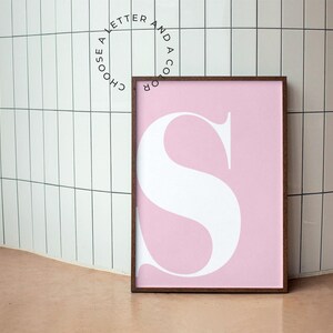 May include: A pink framed print with a white letter "S" in the center. The text around the frame reads "Choose a letter and a color".