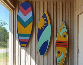 May include: Three colorful surfboards with geometric designs are mounted on a wooden wall. The surfboards feature yellow, blue, teal, pink, and orange hues. The designs include chevrons, suns, and wave patterns, creating a coastal theme.