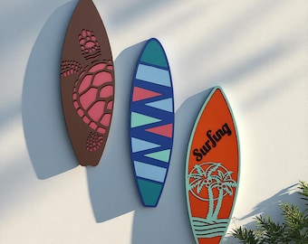 May include: Three decorative surfboards in brown, teal, and orange hang on a white wall. The brown surfboard features a turtle design in pink. The teal surfboard has a blue and pink zigzag pattern. The orange surfboard has the word "Surfing" and palm trees.