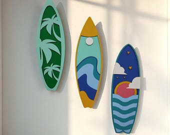 May include: Three decorative surfboards in a row, each with a unique design. The first is green with palm tree cutouts, the second has blue and yellow waves, and the third features a sunset and ocean waves. They are mounted on a white wall.