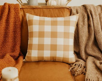 Könnte beinhalten: Gemütliche Wohnzimmerszene mit Herbstdekor. Ein kissen mit tan-weißem Karomuster liegt auf einem braunen Ledersofa, flankiert von orangefarbenen und braunen Decken. Ein kleiner Tisch mit einem weißen Kürbis und einer Kerze.