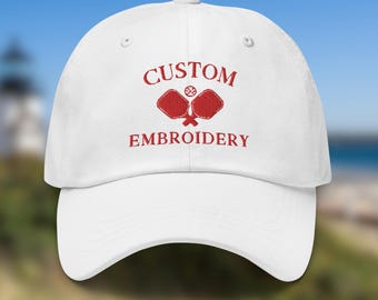 May include: White baseball cap with red embroidery that says "CUSTOM EMBROIDERY" and features a red paddleball and ball.