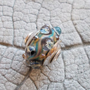 May include: A handcrafted glass bead with a silver-colored metal accent. The bead features a swirling pattern of blue, teal, and brown colors, with clear, bubble-like embellishments. The bead is designed for jewelry making.