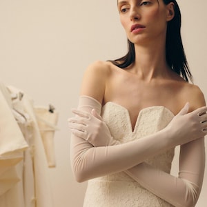 Puede incluir: Un vestido de novia de encaje marfil sin tirantes con escote corazón. El vestido se combina con guantes transparentes hasta el codo. La modelo lleva un delicado tocado. Otros vestidos de novia son visibles en el fondo.