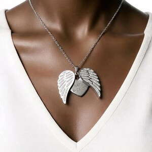 Puede incluir: Collar plateado con un colgante que presenta alas de &aacute;ngel y una etiqueta en forma de coraz&oacute;n. Las alas est&aacute;n adornadas con peque&ntilde;as piedras brillantes. La etiqueta del coraz&oacute;n est&aacute; grabada con el nombre "NATIE" y la fecha "6-28-20".