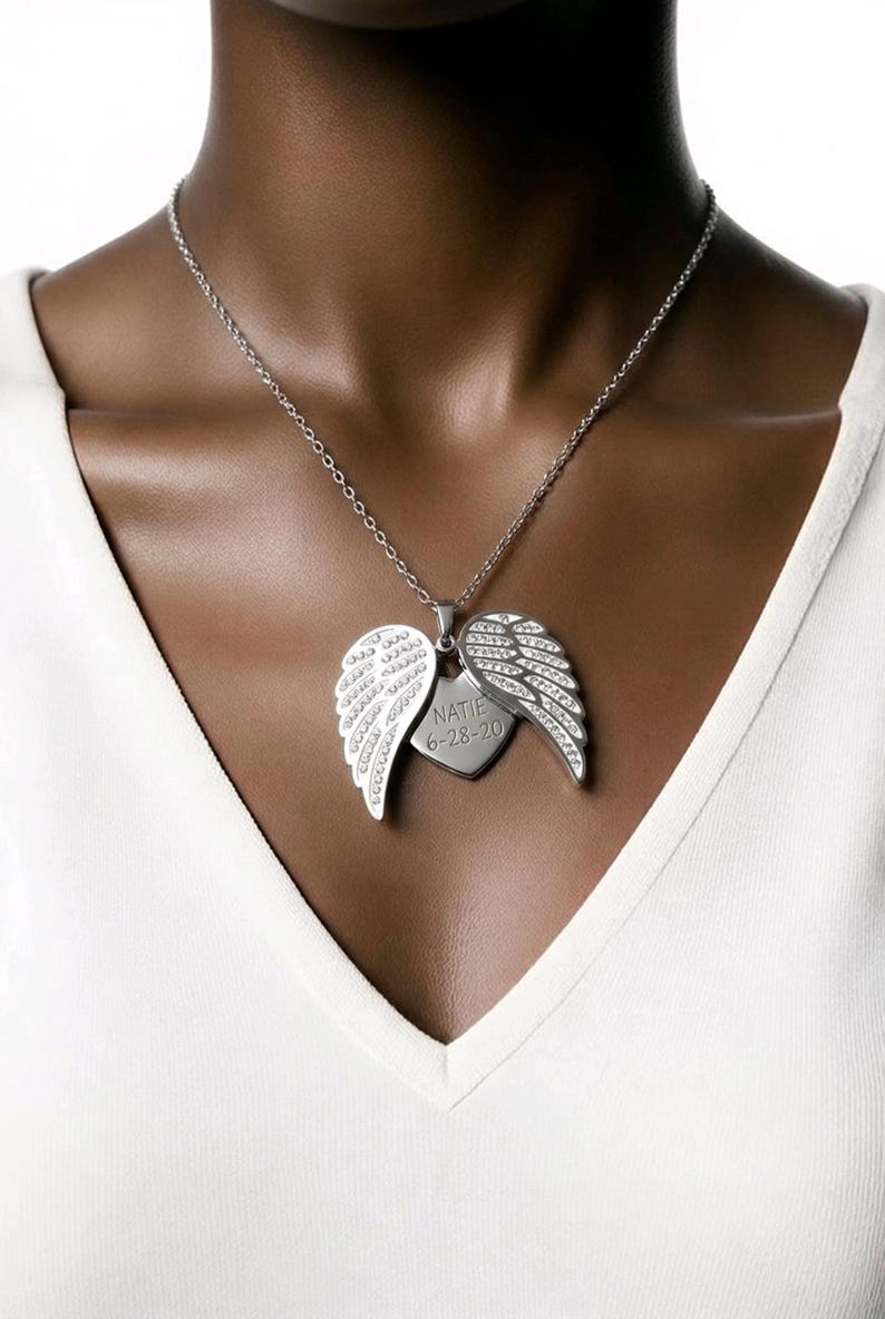 Puede incluir: Collar plateado con un colgante que presenta alas de &aacute;ngel y una etiqueta en forma de coraz&oacute;n. Las alas est&aacute;n adornadas con peque&ntilde;as piedras brillantes. La etiqueta del coraz&oacute;n est&aacute; grabada con el nombre "NATIE" y la fecha "6-28-20".