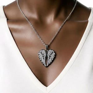 Puede incluir: Collar plateado con colgante en forma de coraz&oacute;n con dise&ntilde;o de alas adornadas con cristales transparentes. El collar se lleva sobre una camiseta blanca con cuello en V. Las alas del colgante son detalladas.