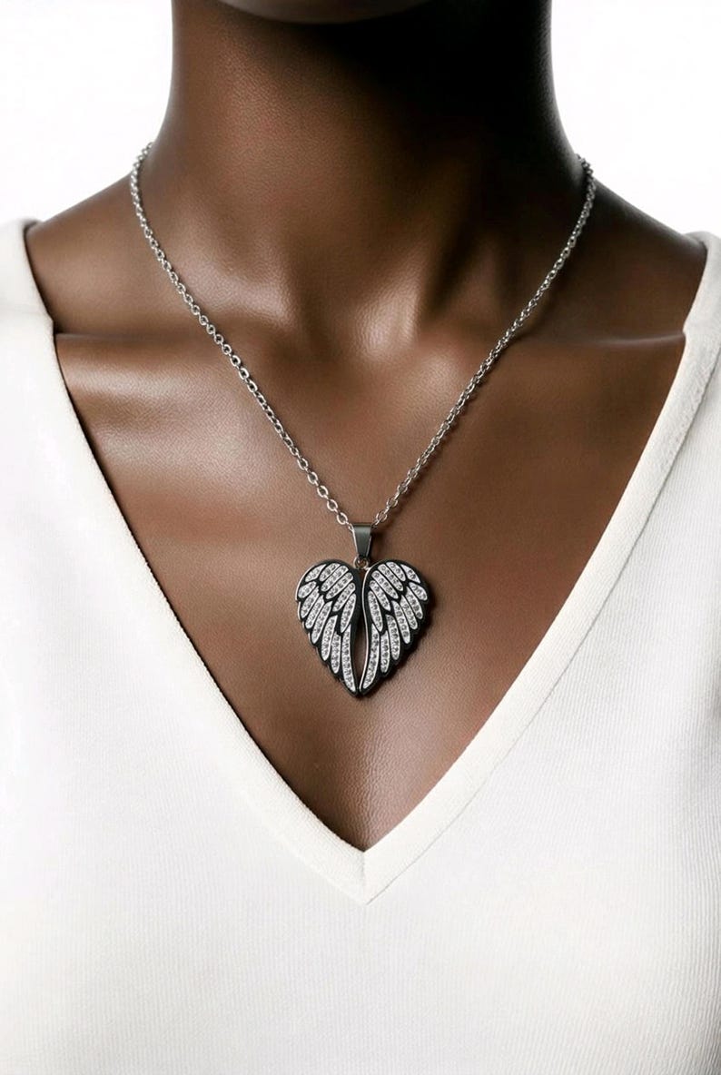 Puede incluir: Collar plateado con colgante en forma de coraz&oacute;n con dise&ntilde;o de alas adornadas con cristales transparentes. El collar se lleva sobre una camiseta blanca con cuello en V. Las alas del colgante son detalladas.
