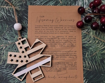May include: Wooden star-shaped ornament with a thin white stripe and stars, resembling a flag, hanging from a string with a white bead. The ornament is on a background with a card that reads "Our Lifesaving Warriors".