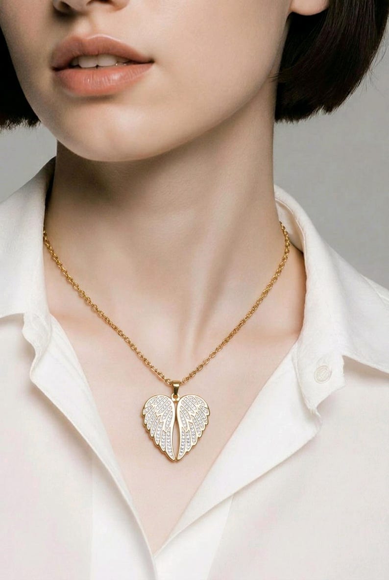 Puede incluir: Un collar dorado con un colgante en forma de alas de &aacute;ngel. Las alas est&aacute;n adornadas con peque&ntilde;os cristales brillantes. El collar se lleva sobre una camisa blanca.