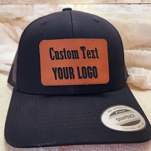 May include: Black trucker hat with a brown leather patch that says "Custom Text YOUR LOGO". The hat has a black mesh back and a black snap closure.