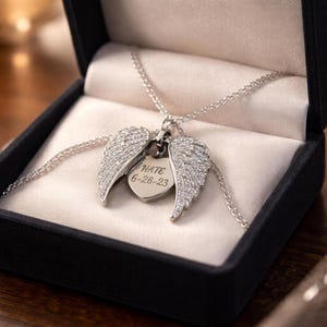 Puede incluir: Un collar plateado con un colgante en forma de coraz&oacute;n y alas de &aacute;ngel abiertas, presentado en una caja de joyer&iacute;a negra. El colgante est&aacute; grabado con el nombre "NATE" y la fecha "6-28-23". Las alas est&aacute;n incrustadas con peque&ntilde;as piedras brillantes.