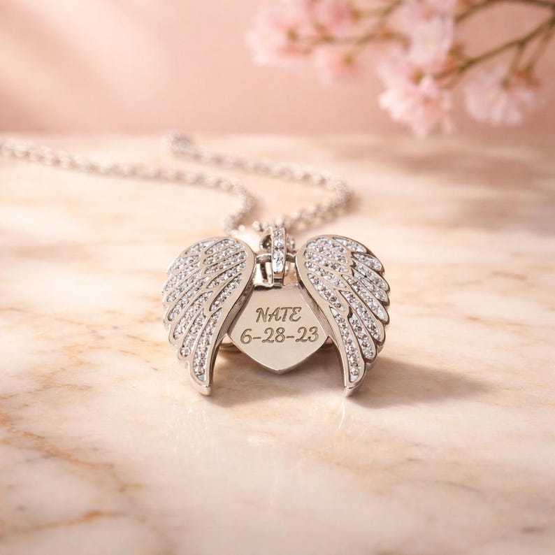 Puede incluir: Collar con colgante plateado con un dije en forma de coraz&oacute;n y dos alas abiertas adornadas con piedras transparentes. El dije tiene grabado "NATE 6-28-23". Se muestra sobre una superficie de m&aacute;rmol con fondo rosa.