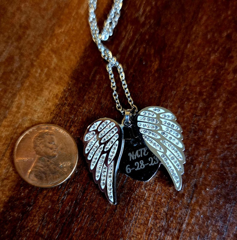 Puede incluir: Collar plateado con un colgante con alas de &aacute;ngel y una etiqueta en forma de coraz&oacute;n. Las alas est&aacute;n adornadas con cristales transparentes. La etiqueta est&aacute; grabada con el nombre "Nate" y la fecha "6-28-23". Una moneda est&aacute; al lado del colgante para la comparaci&oacute;n de tama&ntilde;os.