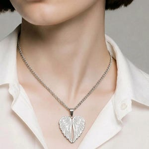 Puede incluir: Un collar plateado con un colgante en forma de alas de &aacute;ngel. Las alas est&aacute;n adornadas con peque&ntilde;os detalles brillantes. El collar cuelga de una fina cadena y se lleva sobre una camisa blanca.