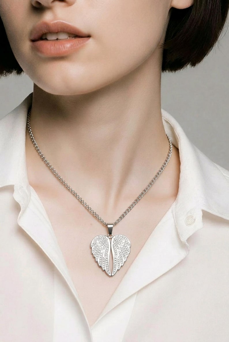 Puede incluir: Un collar plateado con un colgante en forma de alas de &aacute;ngel. Las alas est&aacute;n adornadas con peque&ntilde;os detalles brillantes. El collar cuelga de una fina cadena y se lleva sobre una camisa blanca.