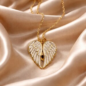 Puede incluir: Un collar dorado con un colgante en forma de alas de &aacute;ngel. Las alas est&aacute;n detalladas con detalles blancos y peque&ntilde;os cristales transparentes. El collar descansa sobre una tela de sat&eacute;n.