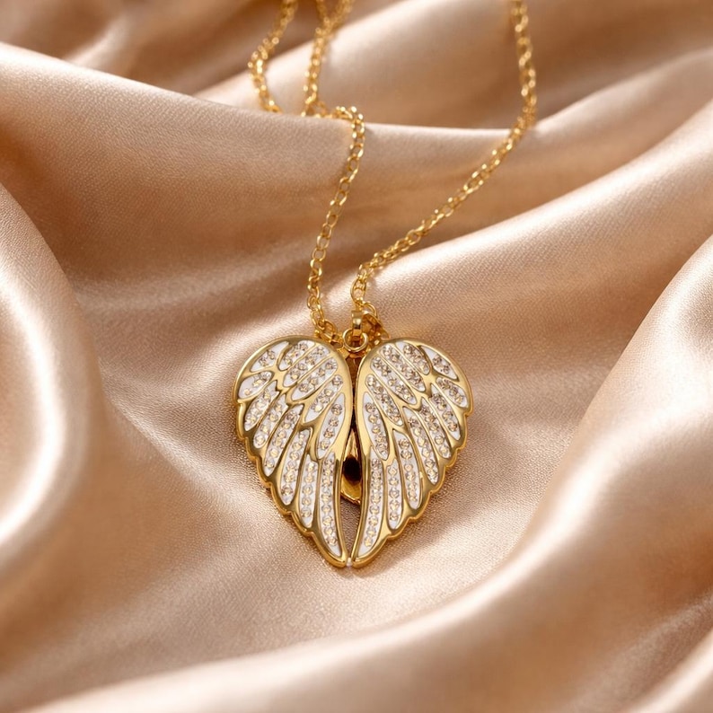 Puede incluir: Un collar dorado con un colgante en forma de alas de &aacute;ngel. Las alas est&aacute;n detalladas con detalles blancos y peque&ntilde;os cristales transparentes. El collar descansa sobre una tela de sat&eacute;n.